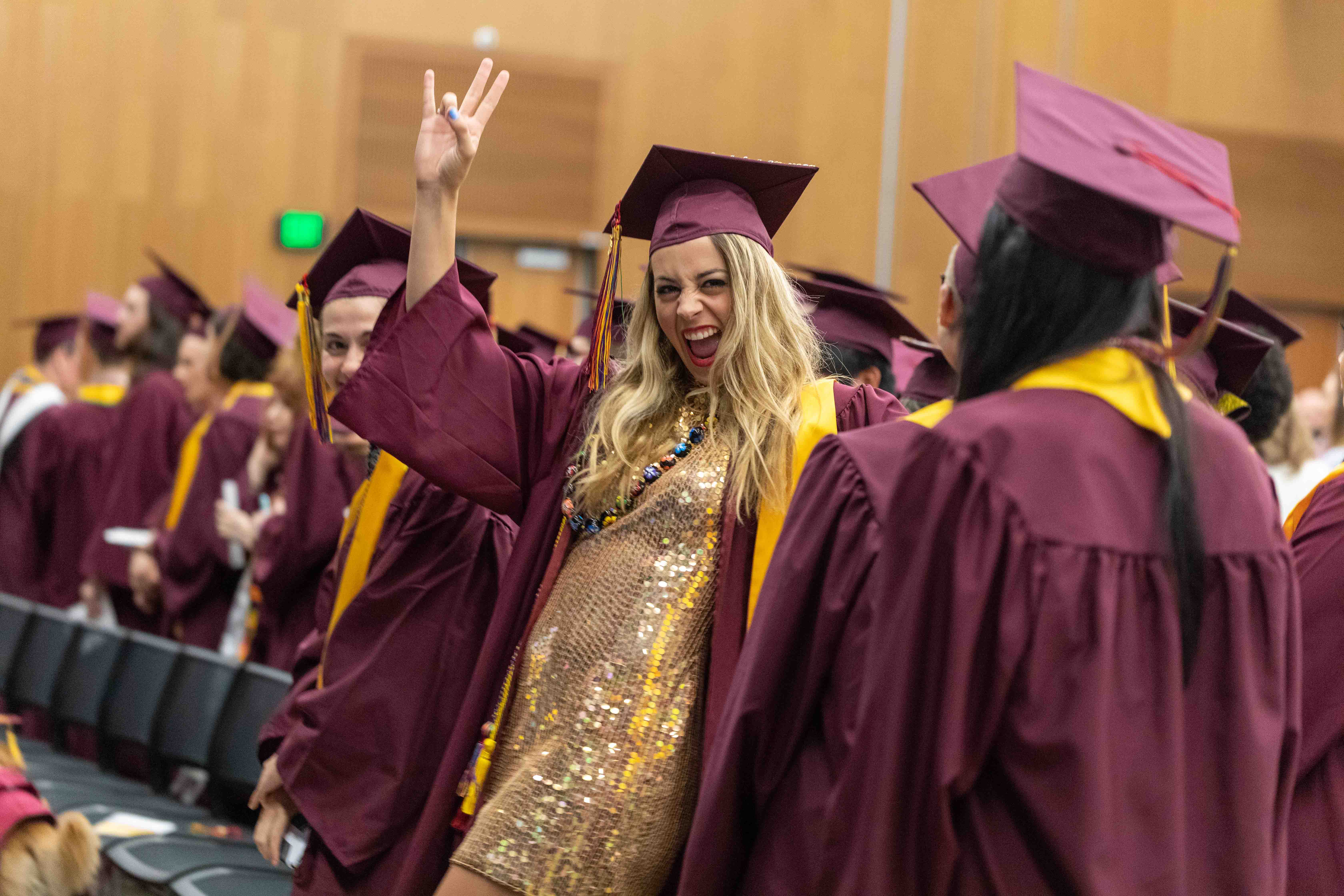 ASU student in a gold sequined dress gives a forks up symbol at the Rainbow LGBTQ graduation convocation at ASU's Tempe campus in the Student Pavilion