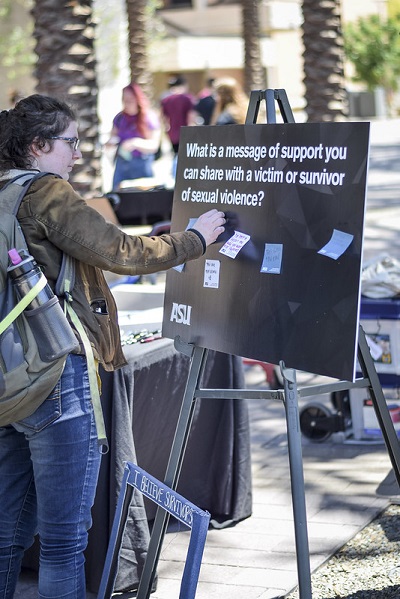 A student places a post-it on a sign that says "what is a message of support you an share with a victim or survivor of sexual violence?"
