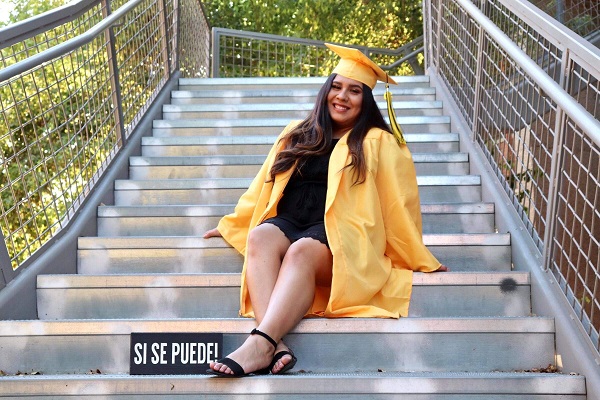 Lorena Mora in her graduation cap and gown on a stairway with a Si Se Puede sign